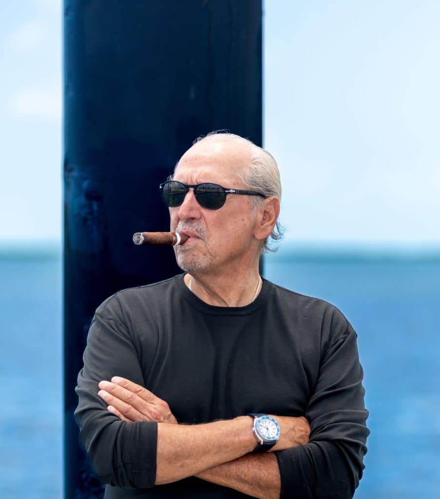 Nestor Miranda standing on a pier facing forward wearing sunglasses and with a cigar in his mouth. Blurred ocean view in the backdrop.