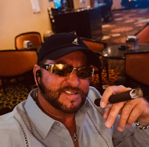 Todd Pribanic seated in a chair in a cigar lounge while wearing sunglasses and holding a lit cigar