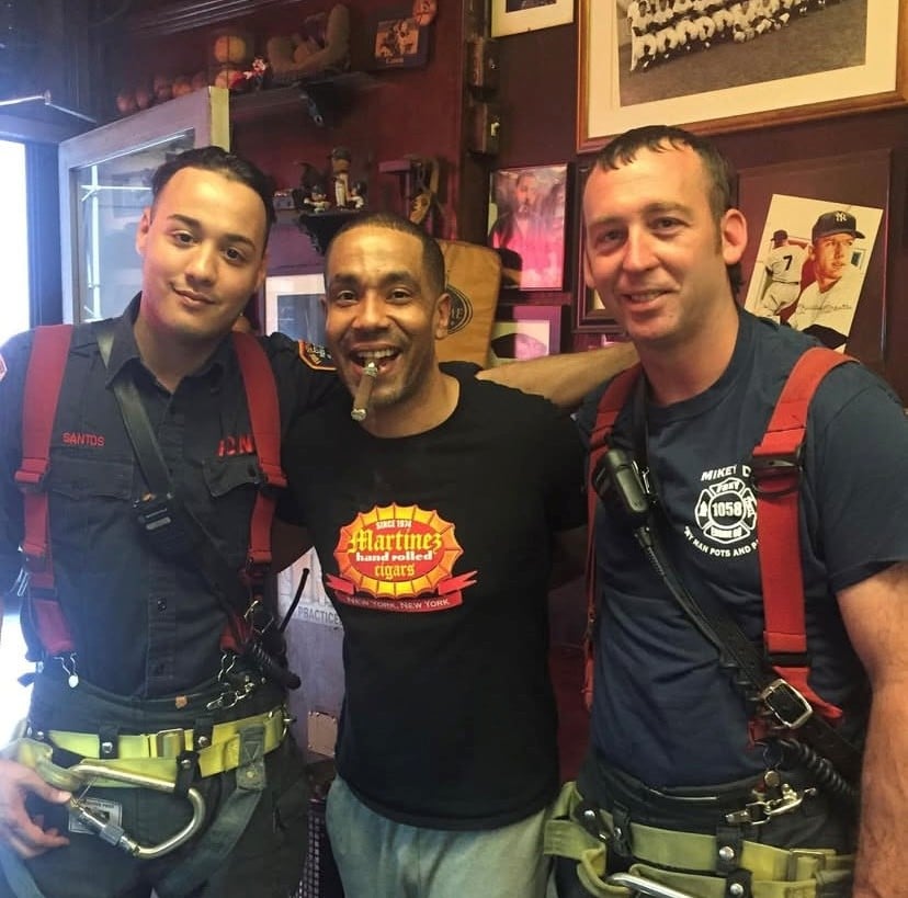 Jesus Martinez standing between two friends while holding a cigar in his mouth