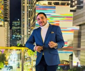 Carlos Nodal wearing a blue blazer and black pants while holding a cigar, the blurred image of a metropolitan city at night is in the backdrop.